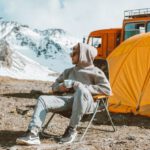 Travel Experiences - Full length of male camper sitting on folding chair next to tent in middle of mountain valley and enjoying views