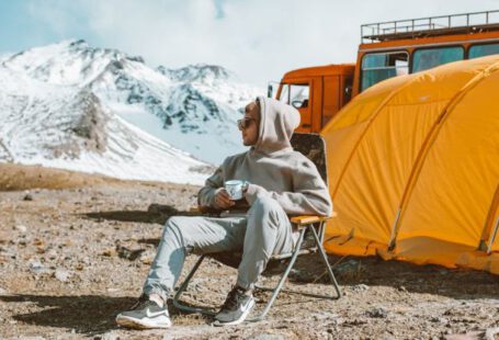 Travel Experiences - Full length of male camper sitting on folding chair next to tent in middle of mountain valley and enjoying views