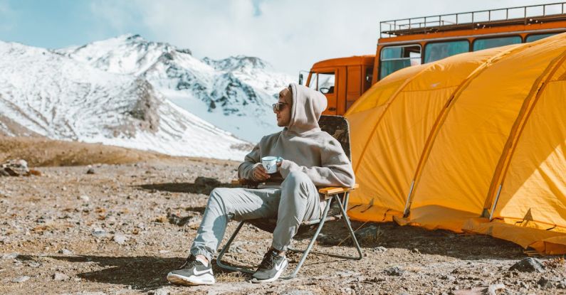 Travel Experiences - Full length of male camper sitting on folding chair next to tent in middle of mountain valley and enjoying views