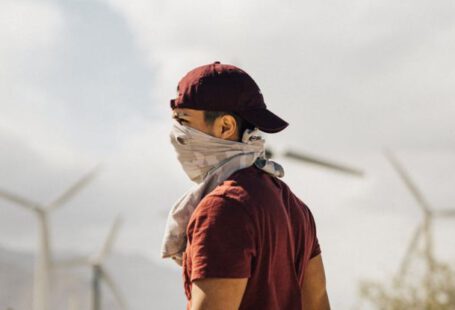 Sustainable Travel - Unrecognizable man with scarf on face walking in sandy terrain terrain near windmills